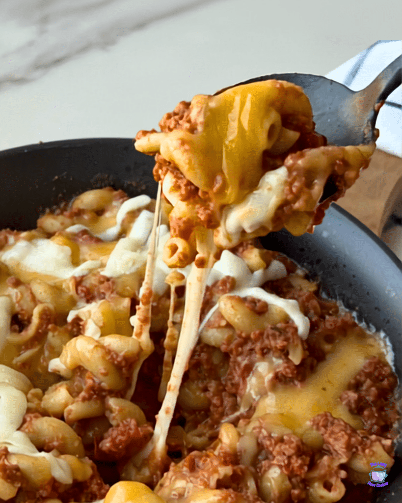 A spoon lifts a serving of cheesy, saucy pasta with ground meat from a skillet, with melted cheese stretching between the spoon and the skillet in this Slow Cooker American Goulash.