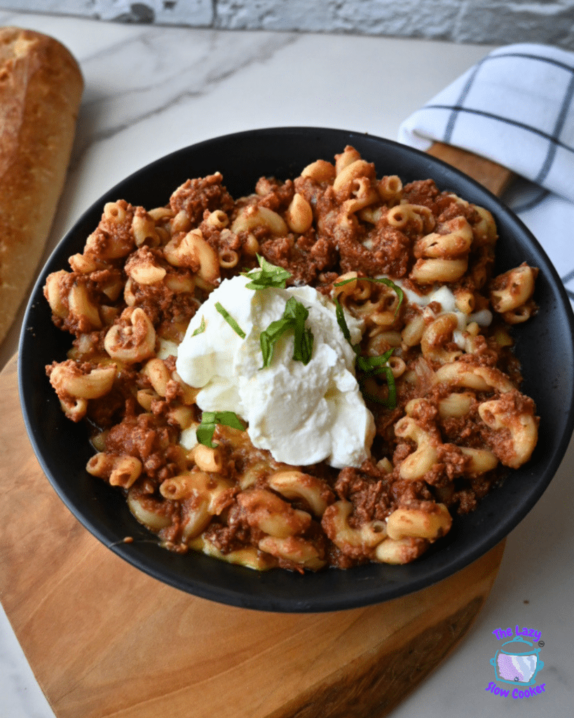 A black bowl holds cheesy beef macaroni, similar to slow cooker American goulash, garnished with a scoop of ricotta and chopped basil. The bowl rests on a wooden board next to a loaf of bread and a folded napkin.