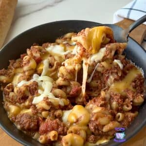 A bowl of cheesy beef macaroni made in a slow cooker is topped with melted cheese. A spoon lifts out a serving, showing the cheese stretching. Slices of bread and a blue-and-white napkin sit in the background.