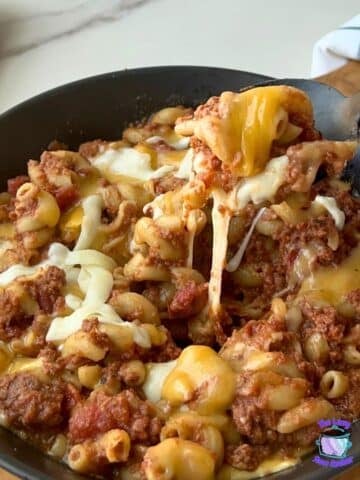 A bowl of cheesy beef macaroni made in a slow cooker is topped with melted cheese. A spoon lifts out a serving, showing the cheese stretching. Slices of bread and a blue-and-white napkin sit in the background.