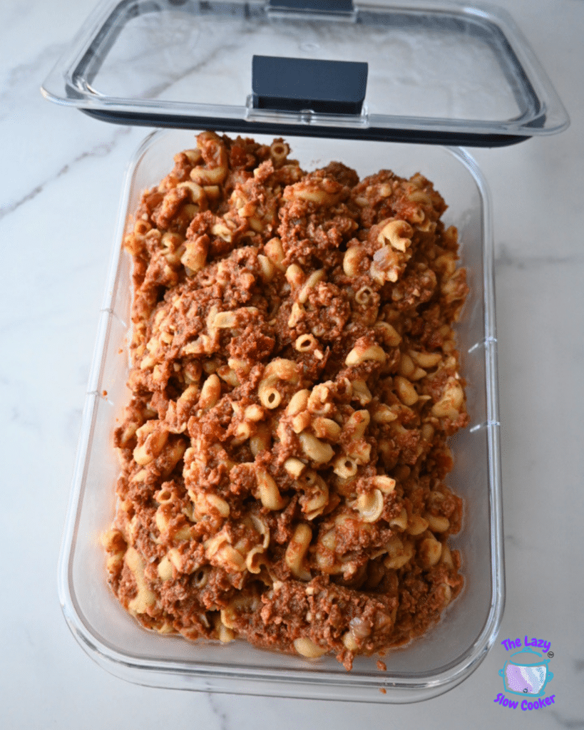 A rectangular container of Slow Cooker American Goulash, showing macaroni pasta mixed with ground meat and tomato sauce, rests on a white surface with the lid set open behind it.
