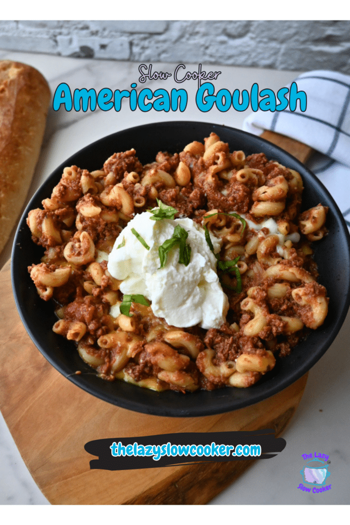 A bowl of American goulash made in a slow cooker is topped with sour cream and fresh herbs, served with a slice of crusty bread. The words “Slow Cooker American Goulash” appear at the top, and the website thelazyslowcooker.com is shown at the bottom.