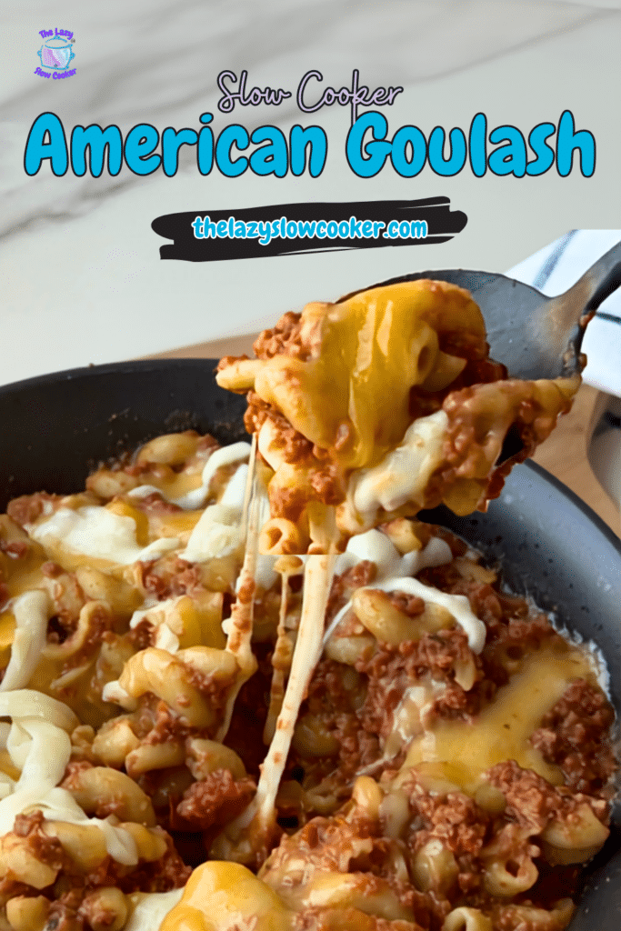 A skillet filled with Slow Cooker American Goulash, made with elbow macaroni and ground meat in a cheesy sauce, is being spooned out. The dish is topped with melted cheese. Text on the image says “Slow Cooker American Goulash thelazyslowcooker.com.”.