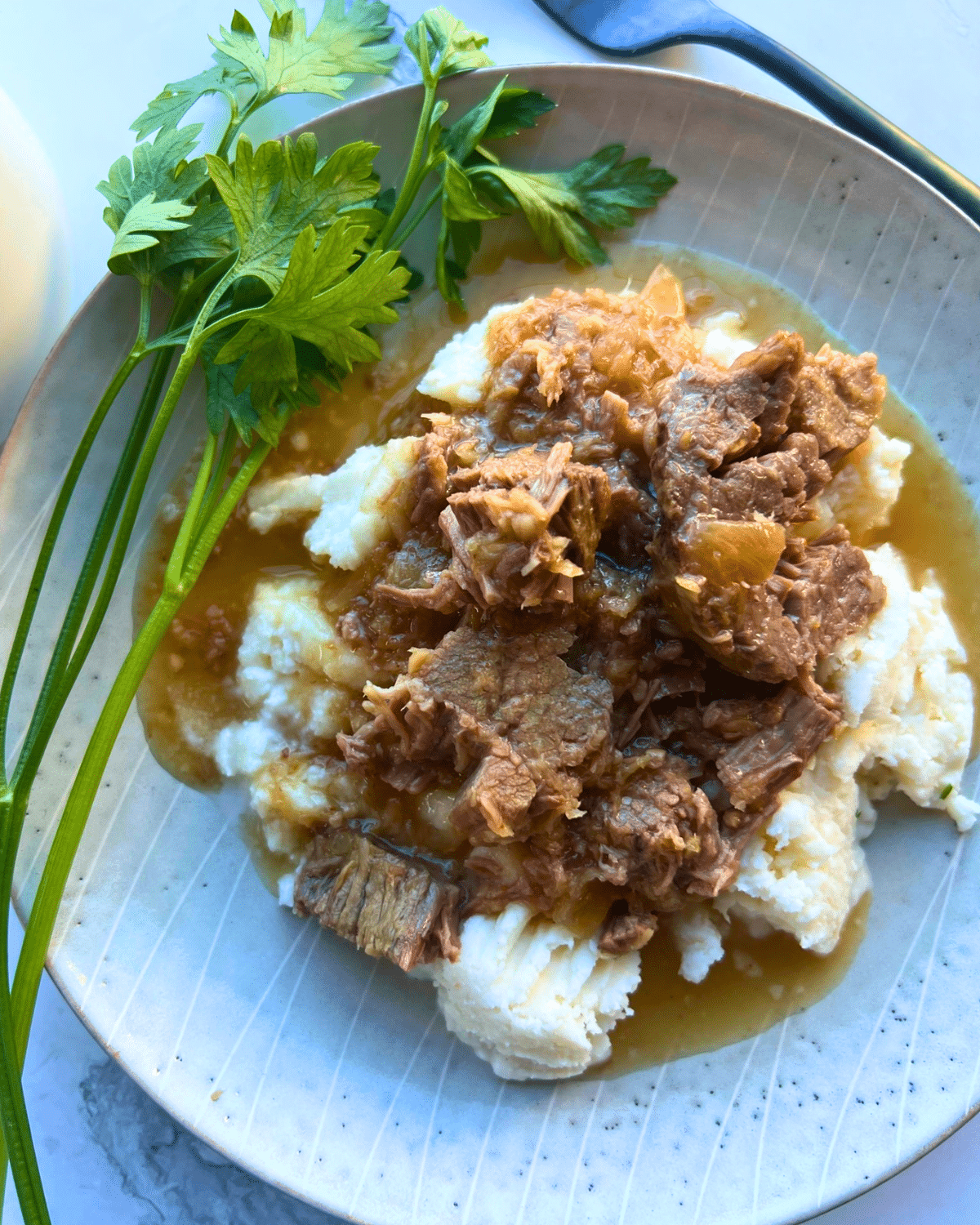 A beige plate holds mashed potatoes topped with tender slow-cooked shredded Horseradish roast and brown gravy, finished with a sprinkle of fresh parsley.