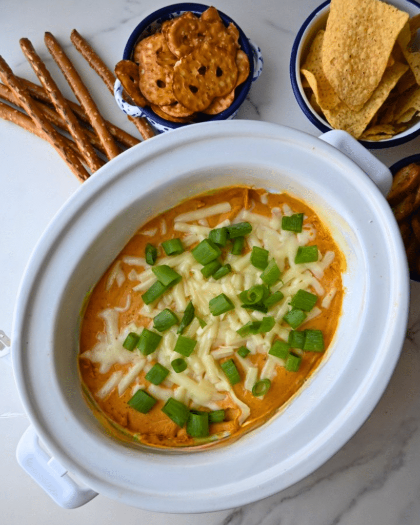 A slow cooker filled with pumpkin dip, garnished with chopped green onions, sits in the center of a white surface. Pretzel sticks, pretzel chips, and tortilla chips are arranged around the slow cooker for dipping.