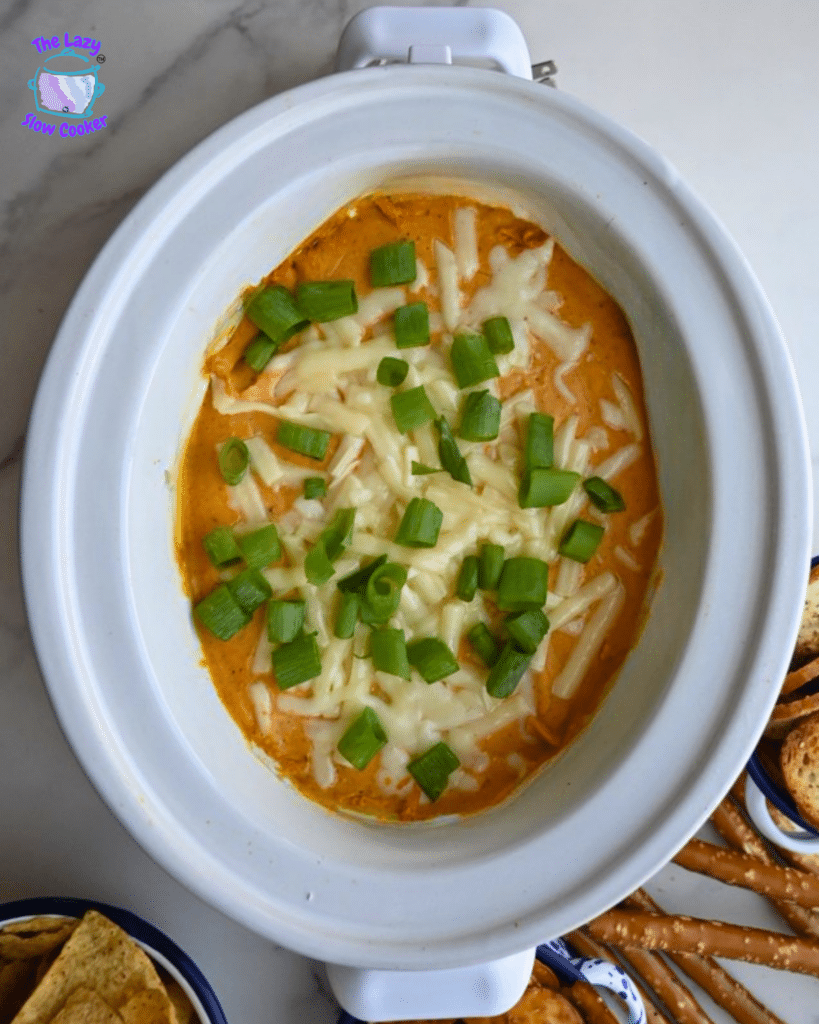 A slow cooker holds creamy pumpkin dip garnished with chopped green onions, set in the center of a white table and surrounded by small bowls of tortilla chips and pretzels for dipping.
