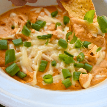 A hand dips a tortilla chip into a white bowl of creamy, cheesy pumpkin dip made in a slow cooker, topped with melted cheese and chopped green onions.
