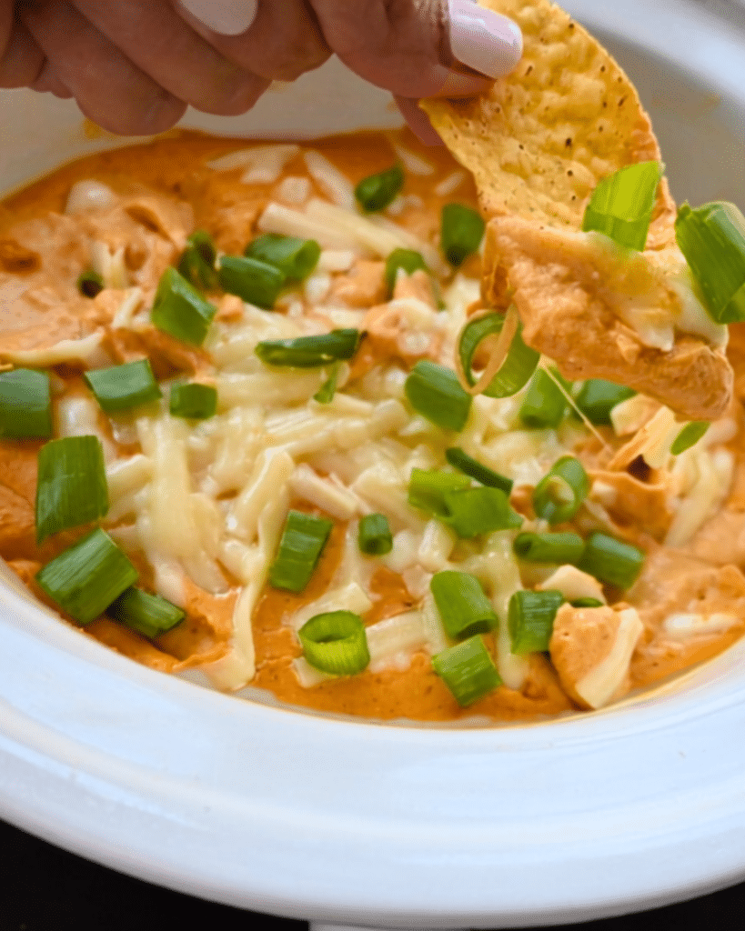 A hand dips a tortilla chip into a white bowl of creamy, cheesy pumpkin dip made in a slow cooker, topped with melted cheese and chopped green onions.
