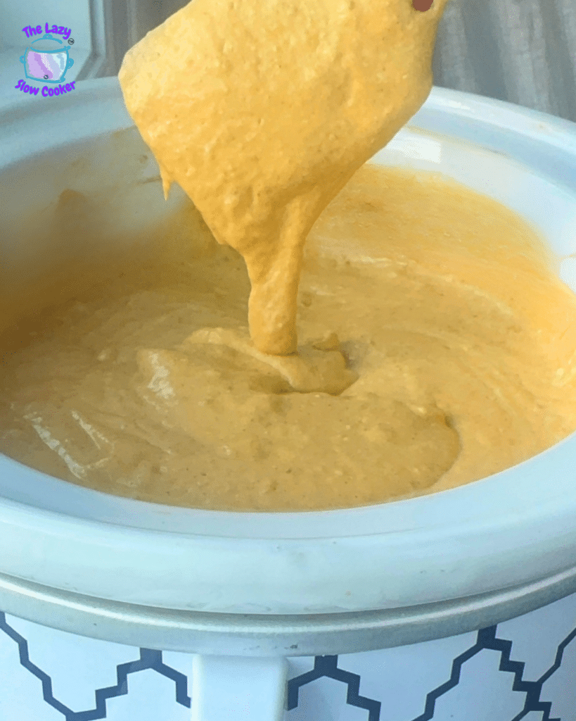 A white slow cooker holds a smooth, creamy yellow mixture being stirred, with a spatula lifting up some of the savory pumpkin dip.