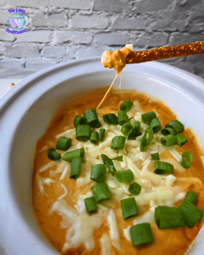 A slow cooker holds pumpkin dip topped with melted cheese and chopped green onions. A hand dips a pretzel into the dip, stretching out gooey cheese. A brick wall is visible in the background.