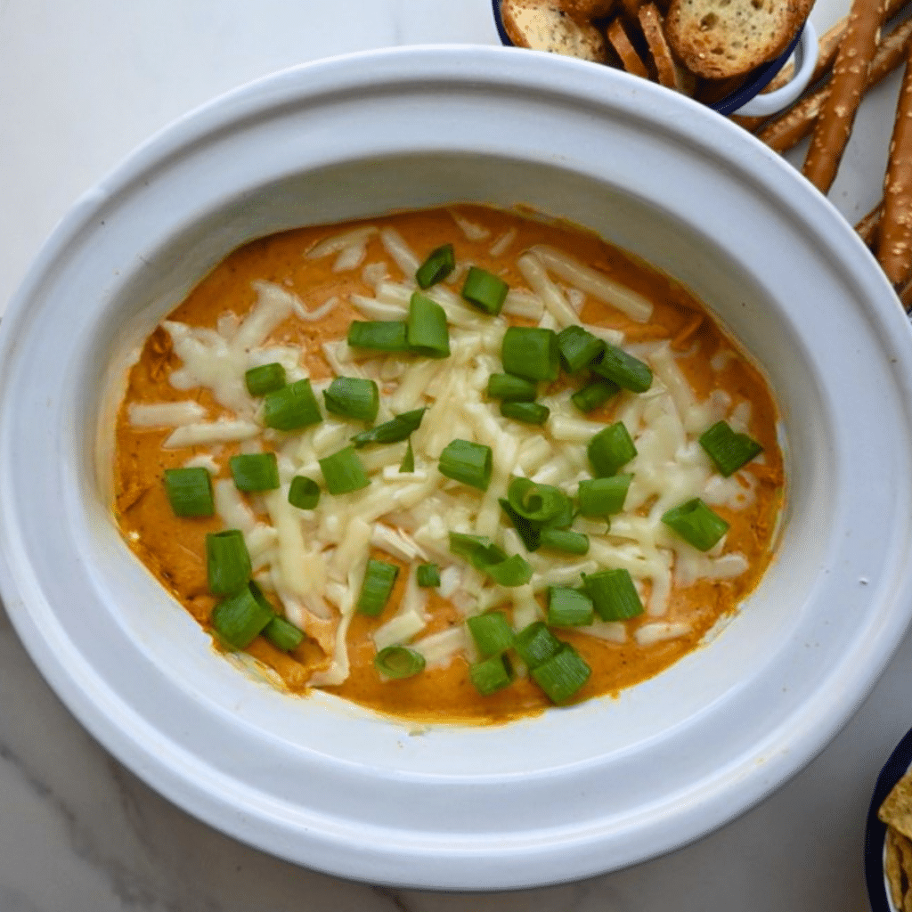 An oval dish holds creamy, savory pumpkin dip made in a slow cooker, topped with melted shredded cheese and chopped green onions. Pretzels and toasted bread slices are arranged along the top edge of the plate.