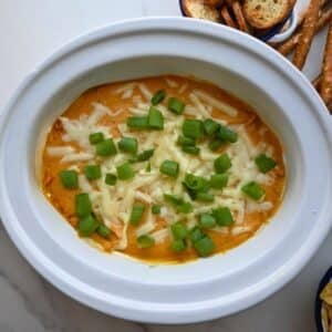 A white dish holds creamy orange pumpkin dip made in a slow cooker, topped with melted cheese and chopped green onions. Pretzel sticks and slices of toasted baguette sit nearby in the background.