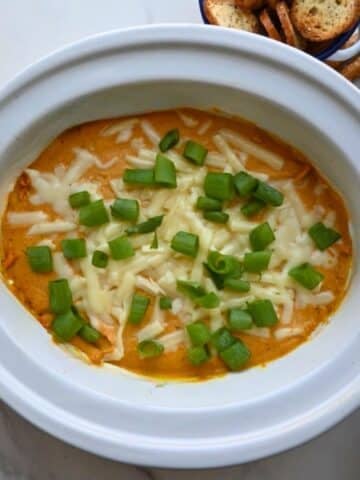 A white dish holds creamy orange pumpkin dip made in a slow cooker, topped with melted cheese and chopped green onions. Pretzel sticks and slices of toasted baguette sit nearby in the background.