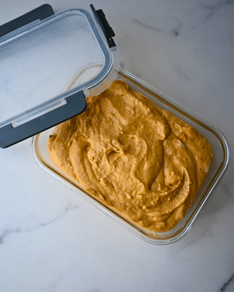 A glass container with a partially open gray plastic lid sits on a white marble surface, filled with creamy orange slow cooker savory pumpkin dip.