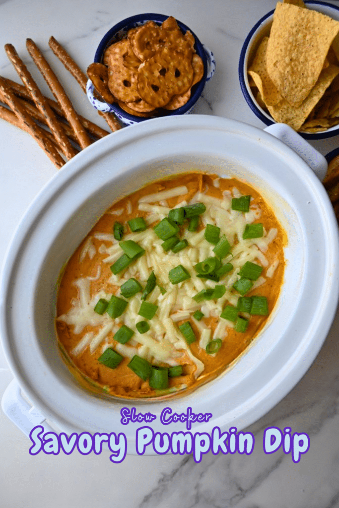 A bowl of savory pumpkin dip made in a slow cooker is topped with melted cheese and sliced green onions. Pretzel sticks, pretzel chips, and tortilla chips are arranged next to the bowl. The image includes the text: Slow Cooker Savory Pumpkin Dip.