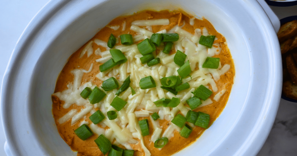 A white dish holds a creamy orange pumpkin dip made in a slow cooker, topped with melted shredded cheese and sprinkled with chopped green onions.