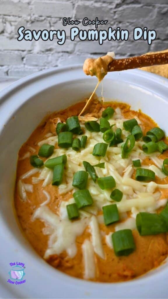 A white slow cooker holds pumpkin dip topped with melted cheese and chopped green onions. A pretzel is being dipped in, and the cheese stretches between the dip and the pretzel. The text on the image says “Slow Cooker Savory Pumpkin Dip.”.