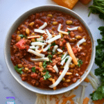 A bowl of hearty chili with edamame, tomatoes, ground turkey, and corn is topped with shredded cheese and set on a marble countertop. Fresh herbs, golden cornbread squares, and extra cheese are arranged around the bowl.